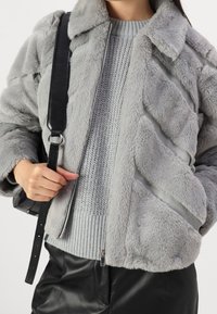Giacca in pelliccia sintetica grigia con motivo a righe diagonali, con colletto e chiusura a zip, indossata sopra un maglione lavorato a maglia grigio chiaro.