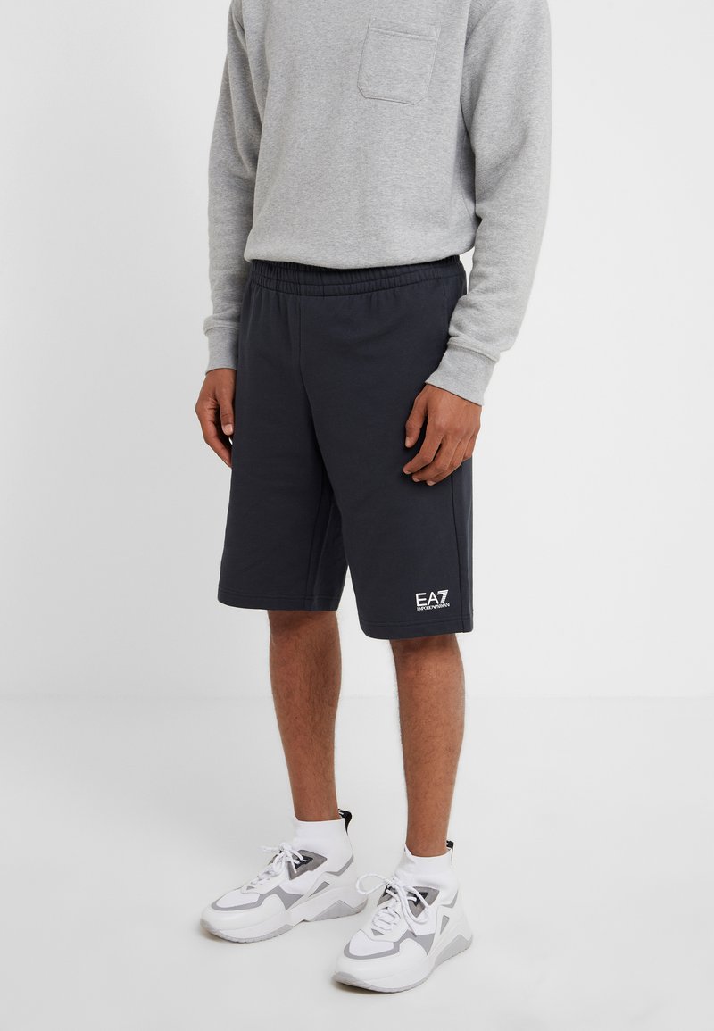 Pantaloni sportivi grigio scuro con elastico in vita, tasche laterali e dettagli con logo. Abbinati a una maglietta a maniche lunghe grigio chiaro e scarpe bianche.
