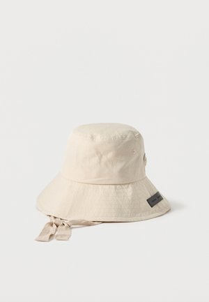 Chapeau bob beige avec large bord et liens en tissu ajustables, comportant de petits trous d'aération latéraux et une étiquette de marque rectangulaire sur le bord.