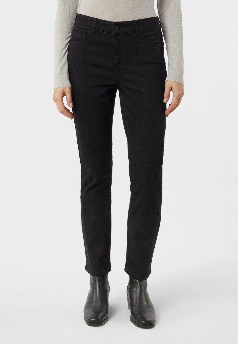 Slim-fit černé džíny v kombinaci s černými kotníkovými botami a světle šedým dlouhým rukávem, zobrazené od půlky trupu po nohy na bílém pozadí.