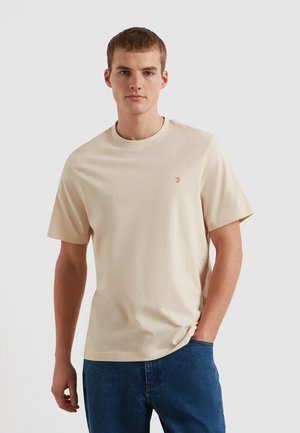 Beżowy bawełniany t-shirt z okrągłym dekoltem, krótkimi rękawami i wyszywanym logo na lewej piersi. Noszony z niebieskimi dżinsami.