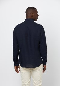 Camicia di lino blu navy con maniche lunghe, polsini a bottone e orlo curvo. Design semplice con una finitura testurizzata, indossata sopra pantaloni di colore chiaro.