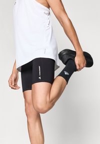 Μαύρο αθλητικό σορτς με εφαρμοστή σχεδίαση, λευκό λογότυπο στο πλάι, συνδυασμένο με λευκό αμάνικο τοπ και μαύρες κάλτσες και παπούτσια κατά τη διάρκεια άσκησης διατάσεων.