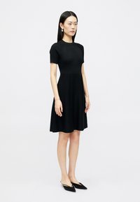 Γυναίκα που στέκεται, φορώντας ένα μαύρο κοντομάνικο ριμπ φόρεμα και μαύρες στιλέτο μπαλαρίνες, σε ένα απλό λευκό φόντο.