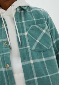 Camicia a quadri turchese con linee bianche, dotata di una tasca frontale, indossata sopra un felpa bianca con cappuccio e cordini.