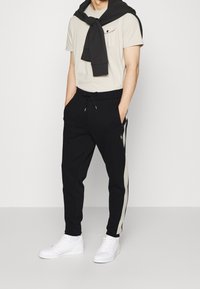 Svarta joggers med vita sidränder, elastisk midja och dragsnöre. Beige t-shirt med korta ärmar och bröstficka. Vita sneakers.