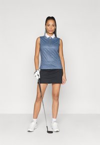 Kék mintás golfpóló fehér gallérral, fekete szoknyával párosítva. A modell golfütőt tart, és fehér sportcipőt visel.