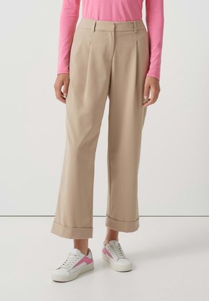 Nainen pukeutuneena beigenvärisiin leveälahkeisiin housuihin, joissa on kalvot, yhdistettynä valkoisiin ja vaaleanpunaisiin tennareihin sekä pitkähihaiseen vaaleanpunaiseen paitaan, seisoo vaalealla lattiapalalla.