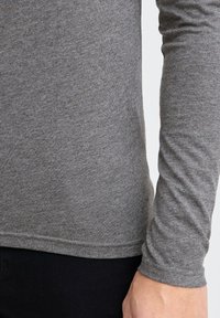 Grå langærmet skjorte lavet af blødt stof med et tætsiddende design. Har en rund sømkant og diskret syning langs ærmerne.