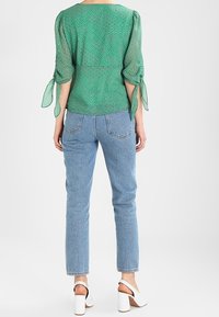 Blouse à motifs verts avec manches bouffantes nouées au poignet, associée à un jean droit bleu clair et des sandales blanches à talons.