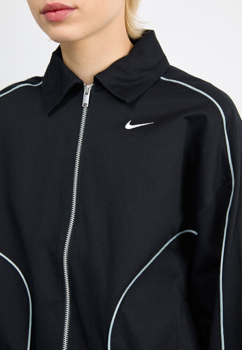 Chaqueta negra de Nike con cierre de cremallera y cuello. Cuenta con un ribete blanco a lo largo de las costuras y el logo de Nike en blanco en el pecho.
