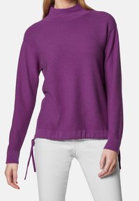 Lila tröja med hög krage, långa ärmar, sidofästen och en texturerad yta. Kombineras med vita jeans för kontrast.