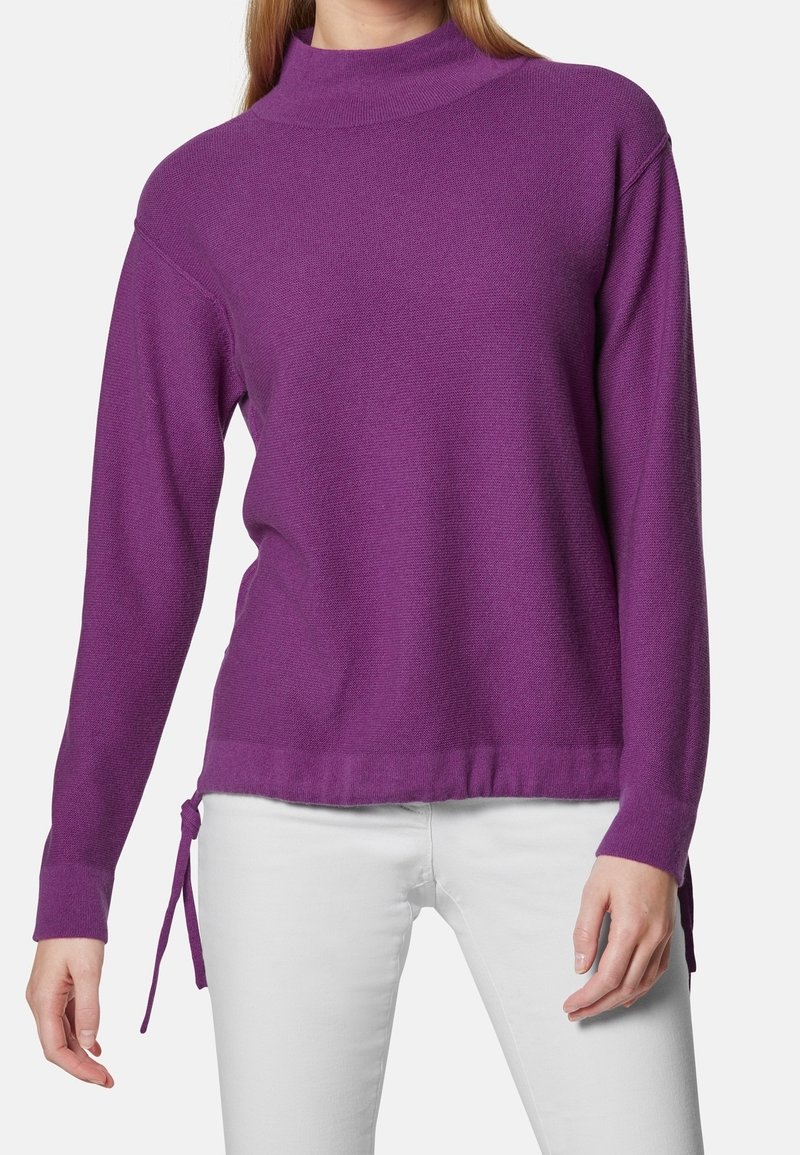 Lila tröja med hög krage, långa ärmar, sidofästen och en texturerad yta. Kombineras med vita jeans för kontrast.