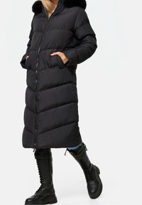 Long manteau matelassé noir avec fermeture éclair, design matelassé horizontal, poches latérales et capuche doublée de fausse fourrure, associé à des bottes noires montantes jusqu'aux genoux.