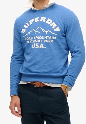 Mand iført en blå sweatshirt med teksten "Superdry Rocky Mountain National Park USA" og bjerggrafik, parret med marineblå bukser og et ur.