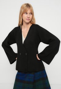 Black knitted jacket with a deep V-neck, double-button closure, and wide sleeves. The model pairs it with a blue and green plaid skirt.