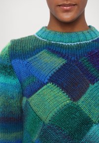 Gros plan sur une personne portant un pull en tricot multicolore avec des motifs losanges bleus, verts et bleu-vert, et un col côtelé.