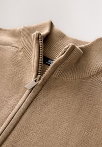 Beige zip-up sweater with ribbed collar and cuffs. Zipper features a plastic pull tab. Soft, textured fabric with a smooth finish.