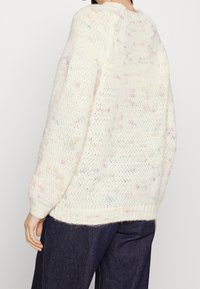 Pull blanc ample en maille avec des taches pastel porté par une personne en pantalon bleu foncé, vu de dos et de côté.