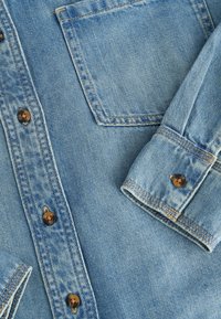 Lichtblauwe denim overhemd met een knoopsluiting aan de voorkant, bruine knopen en een borstzak met zichtbare stiksels en een opgerolde manchette.
