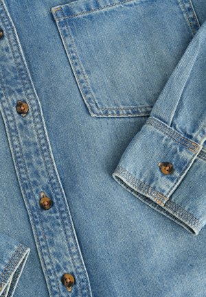 Lyseblå denimskjorte med knaplukning, brune knapper og en brystlomme med synlig syning samt en foldet manchet.