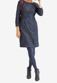 Marineblaues Kleid mit hellblauen Punkten, drei Viertel-Ärmeln, tailliertem Schnitt und knielanger Saum. Kombiniert mit marineblauen Strumpfhosen und Stiefeletten.