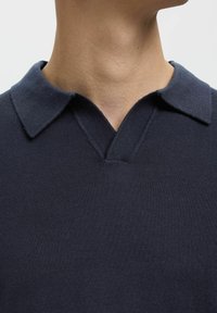 Mornarica pletena polo majica s standardnim ovratnikom, V-izrezom i teksturiranim materijalom. Čiste linije i minimalni detalji.