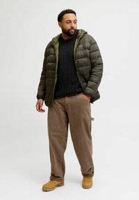 Dark green padded jacket with contrast hood, black cable-knit sweater, beige pants, and yellow boots, standing against a light background.