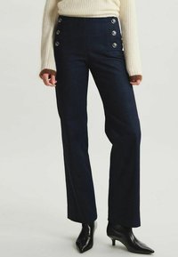 Pantalons en denim foncé taille haute avec une jambe évasée, dotés de boutons en argent audacieux à l'avant et d'une texture lisse.