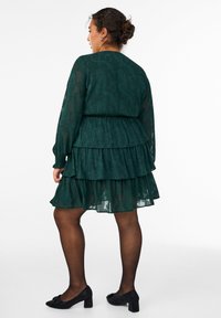 Abito a strati verde realizzato in tessuto trasparente con un motivo floreale, maniche lunghe e vita aderente, abbinato a tacchi neri texturizzati.