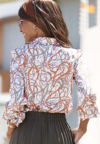 Blusa semitranslúcida con estampado floral, manga con volantes, base blanca y acentos multicolores. Combinada con pantalones plisados oscuros.