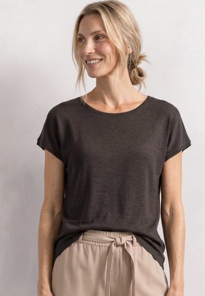 Kvinde med blondt hår i en lav knold iført en mørk kortærmet top og beige bukser med bundet talje, kigger til venstre og smiler.