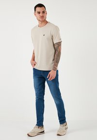 Beige, rövid ujjú póló, kerek nyakkal, kék farmernadrággal és bézs sportcipővel viselve. A pólón kis logó található a mellkason, és a karon tetoválások láthatók.