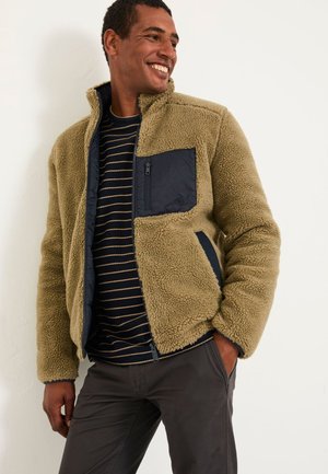 Mann lächelt, trägt eine beige Fleecejacke mit dunkelblauer Brusttasche über einem dunkelblau gestreiften Hemd, steht mit einer Hand in der Tasche vor weißem Hintergrund.