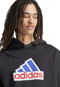 Zwarte hoodie met een groot wit en blauw adidas logo met rode tekst. De stof lijkt glad, en het ontwerp heeft een capuchon en een eenvoudige snit.