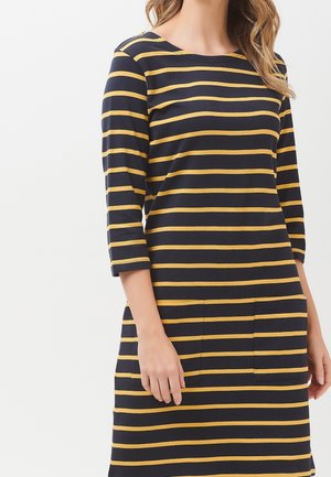 Žena v černých šatech s horizontálními pruhy v barvě hořčicově žluté a tříčtvrtečními rukávy, ruce volně podél těla.