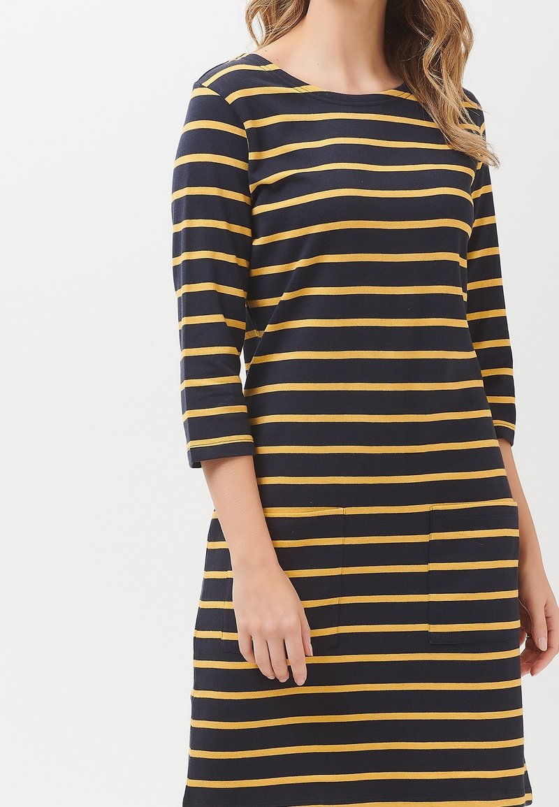 Frau trägt ein schwarzes Kleid mit horizontalen senfgelben Streifen und dreiviertel Ärmeln, die Hände entspannt an den Seiten.