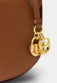 Γωνία καφέ δερμάτινης τσάντας με χρυσά μεταλλικά διακοσμητικά κρεμαστά που είναι συνδεδεμένα σε βρόχο από δερμάτινο ραμμένο λουράκι.