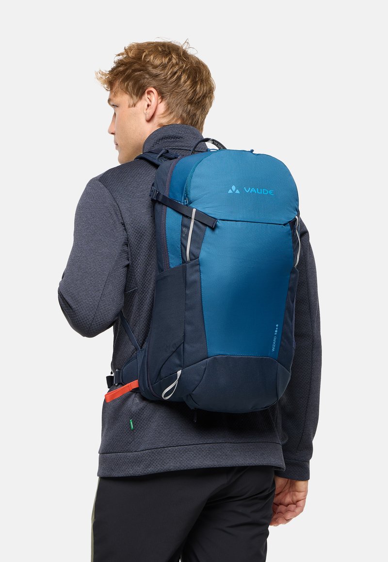 Homme blond en veste sombre portant un sac à dos de randonnée bleu et marine avec des sangles thoracique et ventrale sur un fond uni.