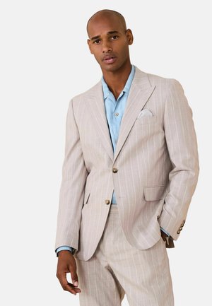 Man wearing beige pinstripe suit with light blue shirt, one hand in pocket, posing against plain white background.