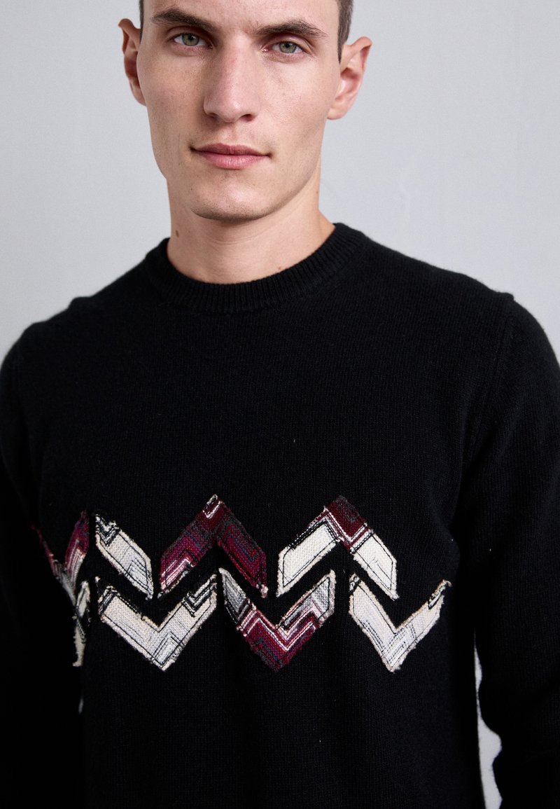 Schwarzer Pullover mit Rundhalsausschnitt, der ein Zickzackmuster in rot, weiß und burgunderrot gestickt über die Brust aufweist.