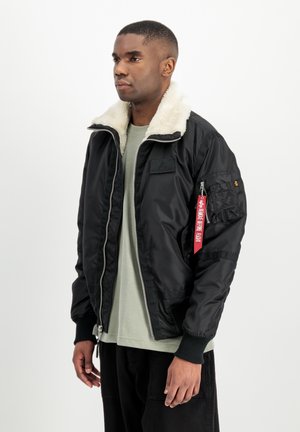 Giovane uomo che indossa una giacca bomber nera con collo in pile bianco e targhetta rossa "Remove Before Flight" sulla manica, in piedi davanti a uno sfondo bianco.