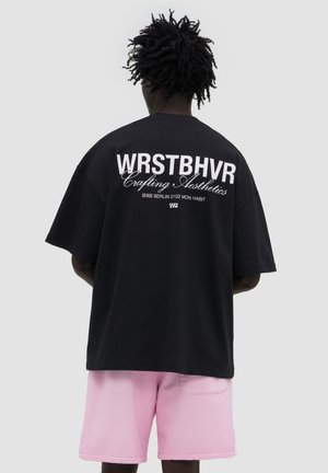 Person mit dunklen lockigen Haaren trägt ein schwarzes übergroßes T-Shirt mit weißem Text und hellrosa Shorts, stehend vor einem einfarbigen grauen Hintergrund.