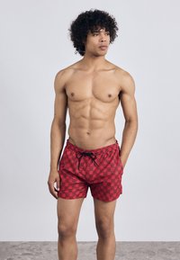 Red swim shorts with a grid pattern featuring "HG" motifs, drawstring waist, and lightweight fabric. Model is shirtless.