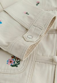 Gros plan sur un vêtement en tissu crème avec un bouton-pression en métal, des coutures cousues et de petites fleurs brodées en rose et bleu.