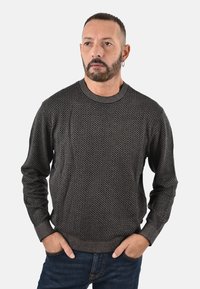 Maglione di maglia grigio con motivo a rilievo, scollatura a girocollo e polsini a coste, abbinato a jeans scuri. Mani infilate nelle tasche.