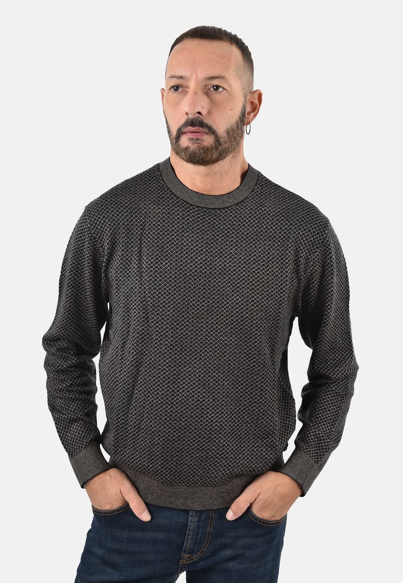 Maglione di maglia grigio con motivo a rilievo, scollatura a girocollo e polsini a coste, abbinato a jeans scuri. Mani infilate nelle tasche.