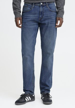 Mann trägt gerade geschnittene blaue Jeans, dunkle Sneakers mit weißen Streifen und einen grauen Langarmpullover, steht vor einem einfarbigen Hintergrund.