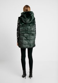 Grüne Fellweste mit Kapuze, ausgestattet mit einem strukturierten Design und horizontalen genähten Paneelen. Kombiniert mit schwarzen Hosen und schnürbaren Ankle-Boots.