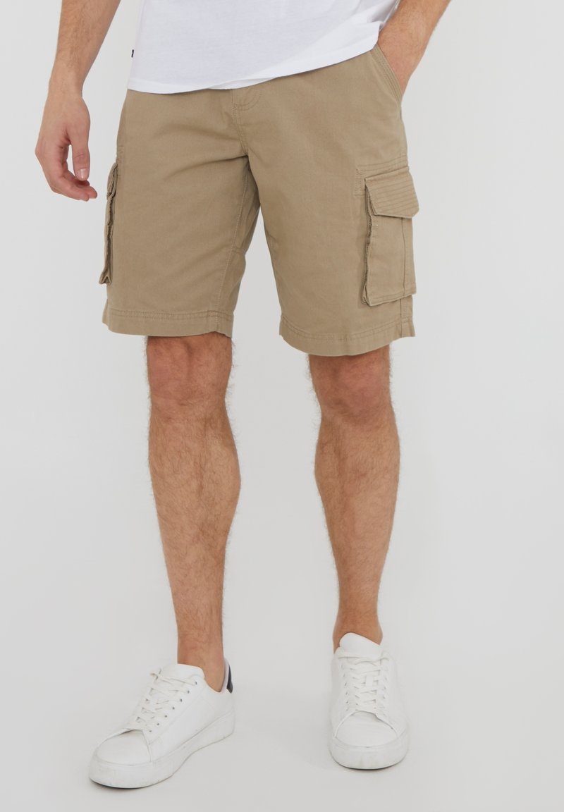 Homme portant un short cargo beige avec des poches latérales et des baskets blanches, debout devant un fond uni.